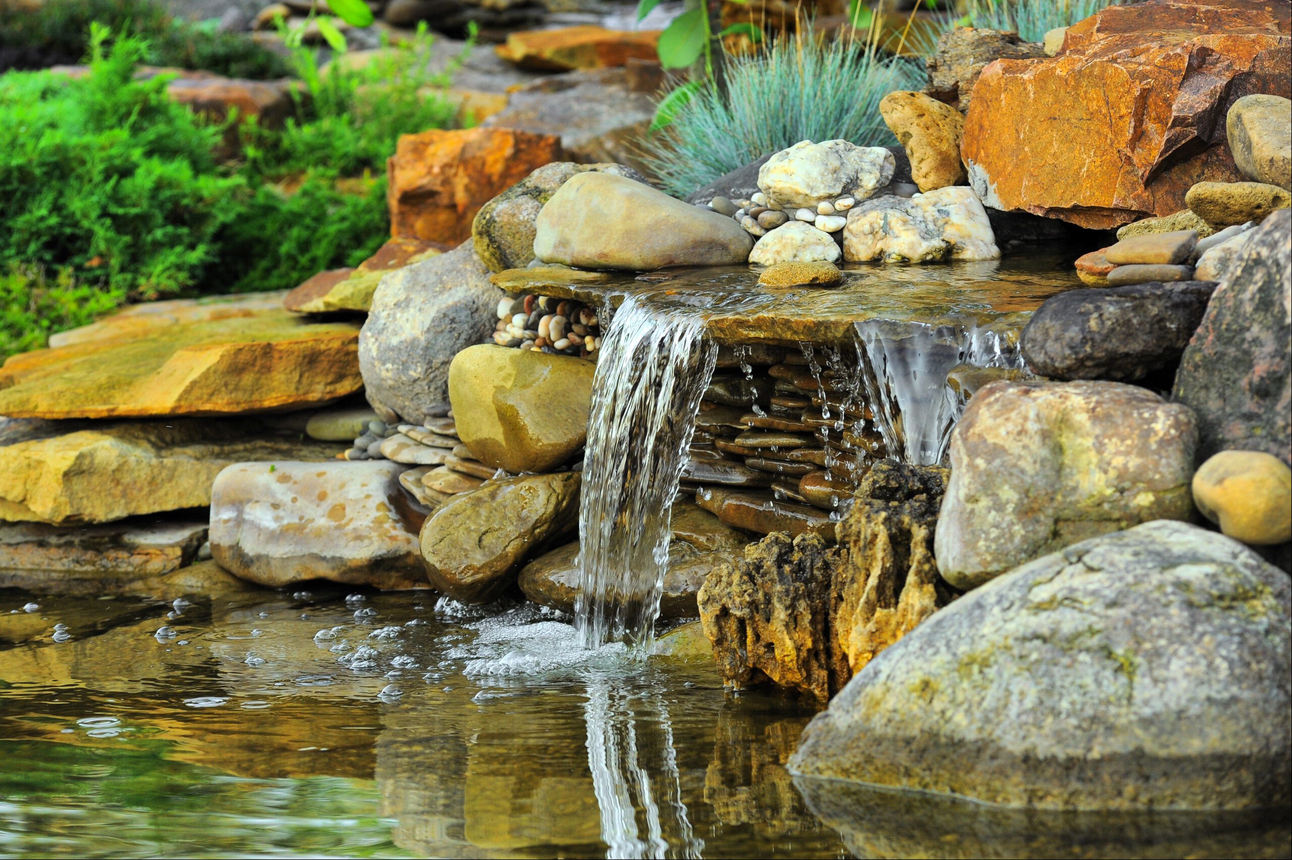 Natural stone waterfall spilling over boulders into a clear pond, surrounded by plantings