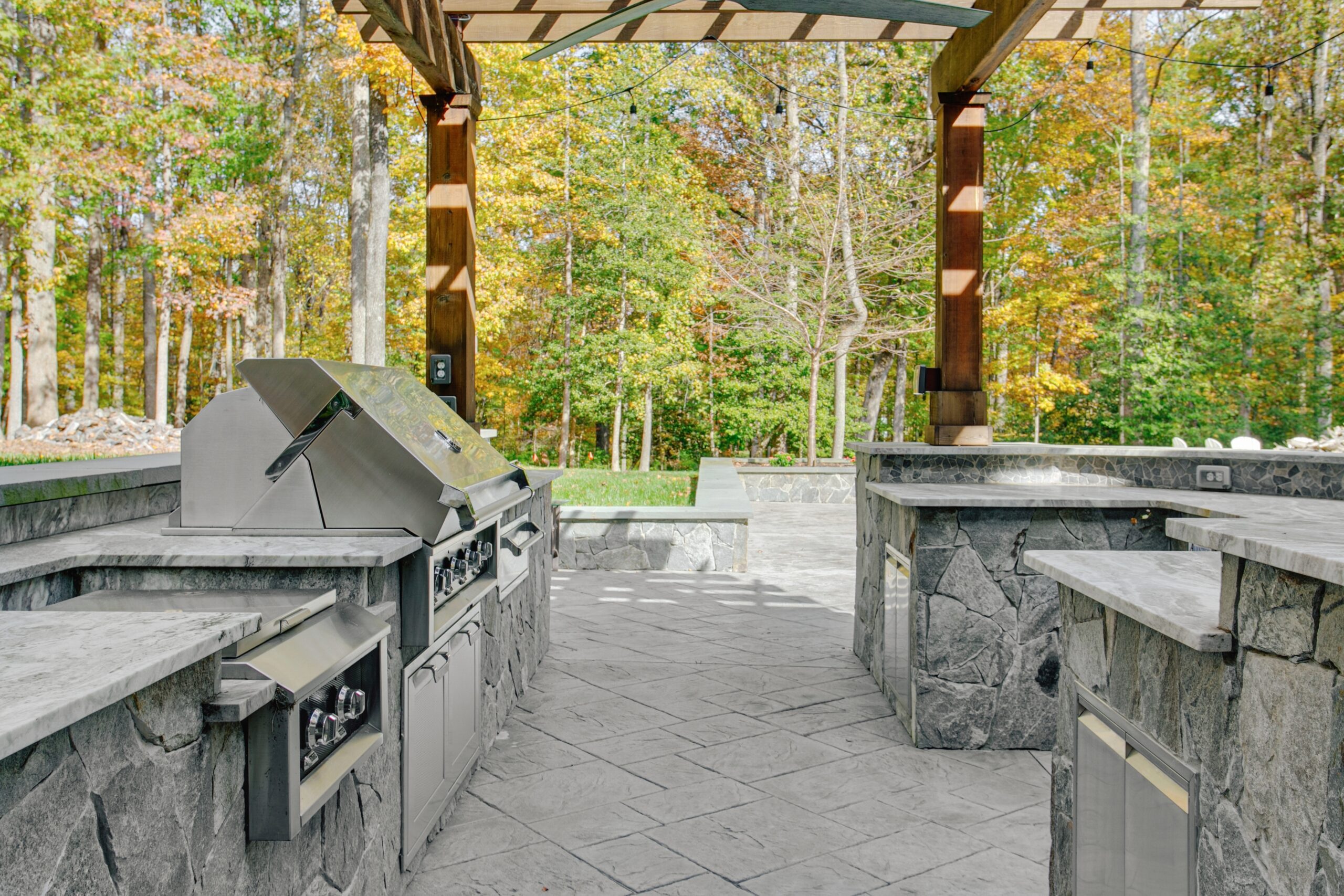 Outdoor kitchen with stone countertops and stainless steel appliances, set under a wooden pergola. Surrounded by a forest with autumn foliage.