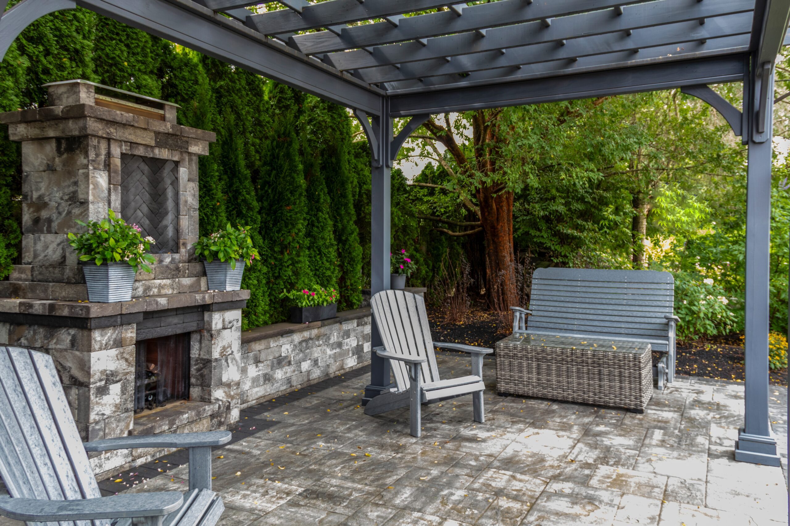 Patio with gray pergola, seating area including chairs and a bench, stone fireplace, and surrounding greenery.